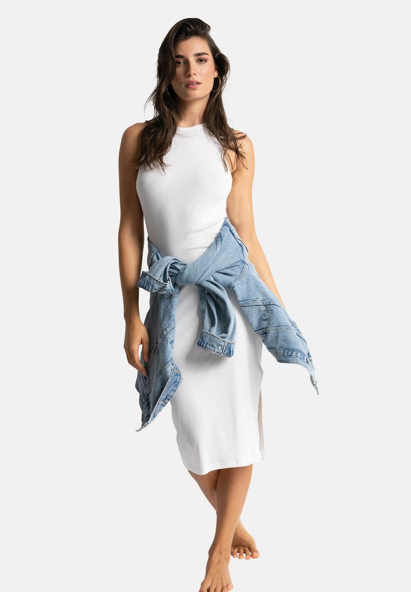 Robe blanche à côtes sans manches avec une fente sur le côté, associée à une veste en denim bleu clair nouée à la taille. Le modèle est pieds nus et se tient debout.