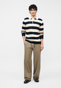 Homme debout portant un polo à manches longues rayé bleu marine et crème, un pantalon large beige et des chaussures marron foncé sur un fond blanc.