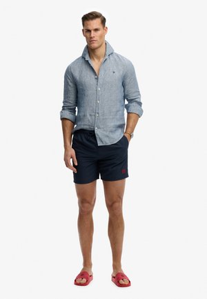 Mand står iført en lyseblå skjorte med knapper og optrukne ærmer, mørke marineblå shorts, røde slidssandaler og et armbåndsur.