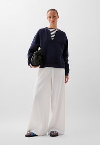 Marineblauwe sweatshirt met een v-hals en geborduurd logo, gelaagd over een gestreept shirt. Gecombineerd met witte bredere broeken en blauwe sandalen. Zwarte tas.