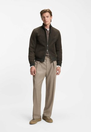 Slim-fit, mörkbrun läderbomberjacka med hög krage, dragkedja och ribbade ärmslut. Beige ulltröja, ljusbeiga byxor och sneakers.