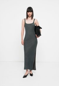 Denim maxi dress in dark gray with a fitted bodice, wide straps, and side slit. Paired with a black clutch and heeled shoes.