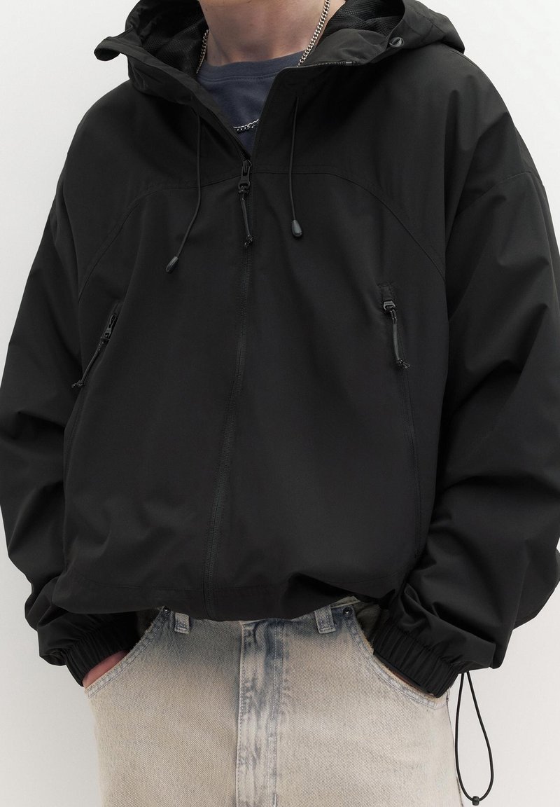 Personne portant une veste coupe-vent noire à capuche avec des poches zippées et un jean bleu clair délavé, les mains dans les poches.