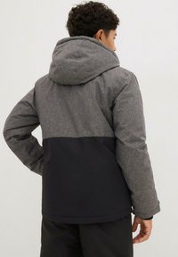 Veste à capuche grise et noire avec une section supérieure texturée et une section inférieure lisse, présentant une coupe décontractée et des détails de couture visibles.