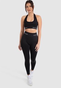 Schwarzes Sportset mit einem Neckholder-Crop-Top mit gebrandetem Band und hoch taillierten Leggings, beide aus einem glatten, dehnbaren Material.