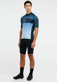 Maillot de ciclismo de manga corta, color azul degradado, hombros texturizados, cierre con cremallera. Combinado con pantalones cortos de ciclismo negro y zapatillas.