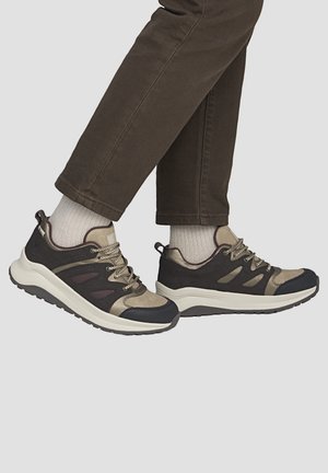 Eine Person, die dunkelbraune Hose, weiße Socken und schwarz-beige Schnürsneaker mit weißen Sohlen trägt und vor einem einfarbigen Hintergrund steht.