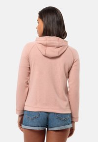 Felpa texturizzata rosa chiaro con cappuccio, caratterizzata da un design aderente e maniche lunghe, abbinata a pantaloncini di denim. Collo alto, senza hardware visibile.