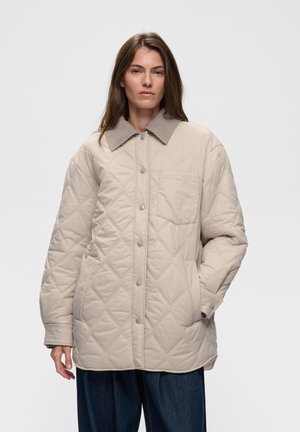 Femme portant une veste matelassée beige avec un col en velours côtelé et un pantalon bleu foncé, debout devant un fond clair uni.