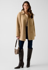 Cappotto di lana beige con colletto a risvolto, quattro bottoni e tasche laterali; abbinato a jeans scuri, stivali di pelle marrone al ginocchio e una borsa a tracolla con stampa leopardata.
