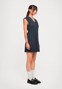 Kvinne med tatoveringer iført en ermeløs marineblå kjole, hvite sokker og svarte loafers, stående mot en enkel lys bakgrunn.