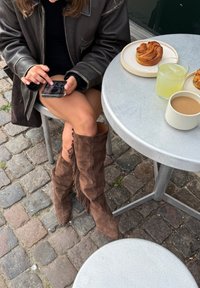 Botas de ante marrón hasta la rodilla con detalles de flecos; usando un vestido negro, sentada en una mesa de metal con pasteles y bebidas. Teléfono en la mano.