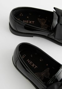 Zwarte lakleren schoenen met een ronde neus en een satijnen strik aan de voorkant. Het interieur heeft een vlinderontwerp met de merknaam "NEXT."