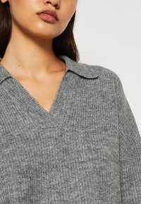 Grå stickad tröja med V-ringning och krage. Texturerad tyg med ett subtilt mönster, med avslappnad passform och mjuk fall.