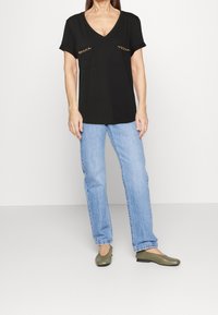 Camiseta negra de cuello en V con mangas cortas, que presenta dos bolsillos delanteros con detalles en cadena dorada, combinada con jeans rectos de tiro claro y bailarinas verdes.
