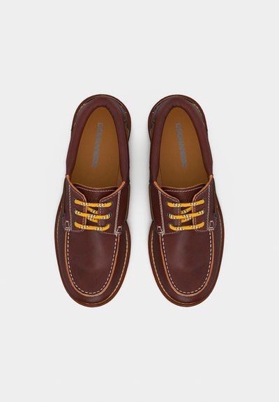 Chaussures bateau en cuir marron avec lacets jaunes et noirs, coutures blanches contrastantes et col rembourré, vues de dessus sur un fond blanc.