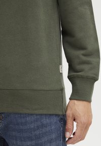 Olivengrønn hettegenser med ribbestrikkede mansjetter og nederkant. Stoffet ser ut til å være teksturert. Liten etikett med tekst plassert på undersiden. Blå denimjeans synlige.