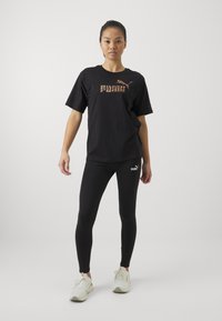 Černé tričko s oranžovým vzorovaným logem, spárované s černými legínami a světle zbarvenými sportovními botami. Jednoduchý, přiléhavý design.