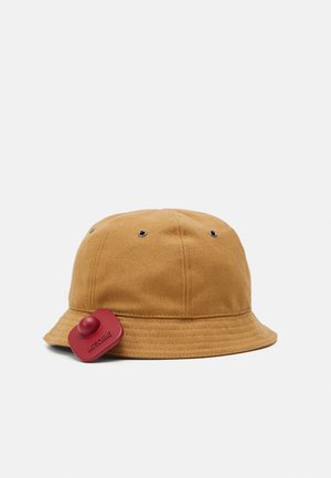 Chapeau seau en coton brun clair avec une finition texturée, comportant des œillets en métal argenté et une étiquette en caoutchouc rouge avec le nom de la marque sur le côté.