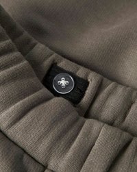 Regulowany elastyczny pasek w talii na szarym materiale z przezroczystym plastikowym guzikiem i czarną listwą na guziki.