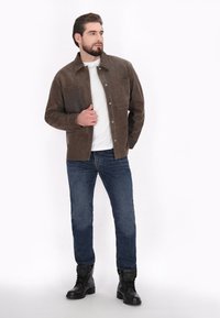 Veste en daim marron avec deux poches poitrine, t-shirt blanc, jean bleu et bottes de combat noires, se tenant contre un fond clair.