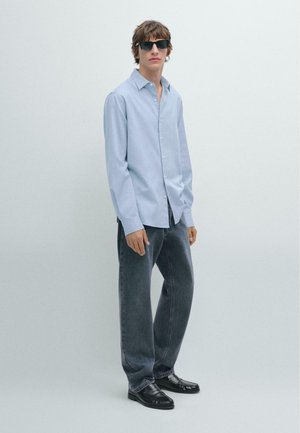 Camicia azzurra chiara a maniche lunghe, jeans scuri grigi a taglio morbido e mocassini neri. Il modello indossa occhiali da sole neri. Tessuto morbido, design casual.