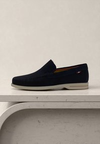 Μπλε σκούρο σουέτ loafer με λευκή σόλα σε φωτεινή πέτρινη επιφάνεια, με μια μικρή κόκκινη, λευκή και μπλε υφασμάτινη λεπτομέρεια κοντά στην φτέρνα.