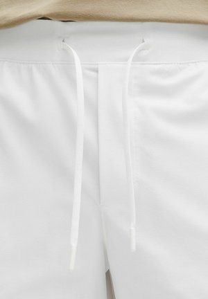 Valkoiset shortsit, joissa on beige joustava vyötärönauha ja kaksi pitkää valkoista kiristysnauhaa roikkumassa keskellä edessä.