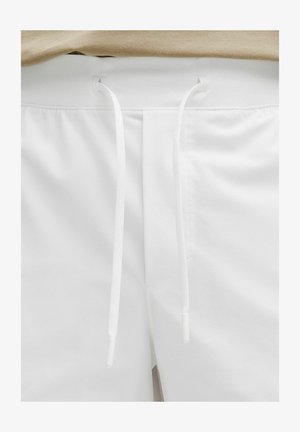 Valkoiset shortsit, joissa on beige joustava vyötärönauha ja kaksi pitkää valkoista kiristysnauhaa roikkumassa keskellä edessä.
