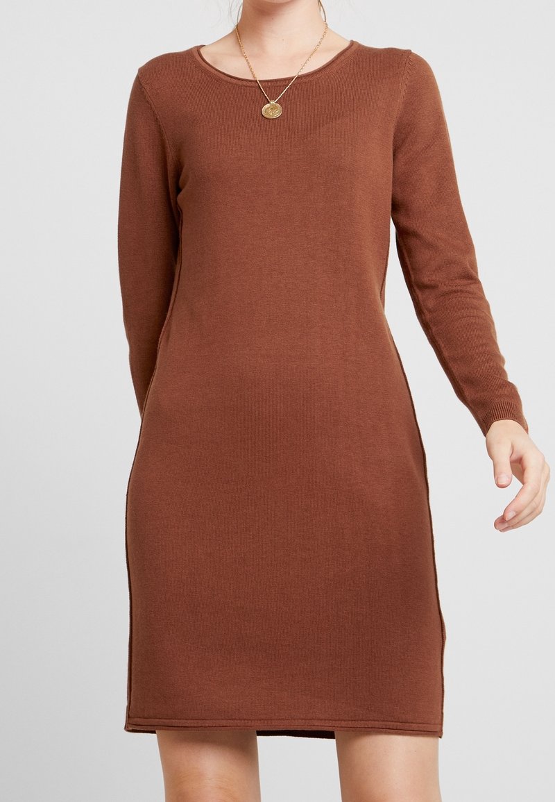 Femme portant une robe marron à manches longues en tricot avec un col rond et un collier pendentif en or sur un fond clair uni.