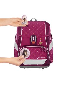 Sac à dos violet avec un motif en diamant et des accents gris ; présente des étiquettes suspendues, des patches et des graphiques de chevaux ; une main attachant un patch rond.