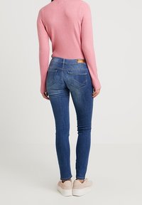 Slim jeans i mörkblå denim, med bakfickor och en beige etikett i midjan. Kombinerat med en ljusrosa tröja och beige sneakers.