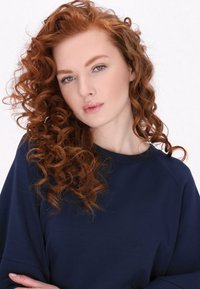 Modella con capelli rossi mossi e lunghi fino alle spalle, che indossa un top blu navy a maniche lunghe con scollatura tonda. Texture liscia, dettagli minimi.