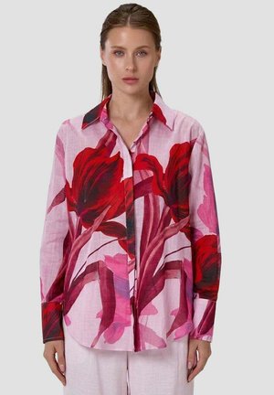 Femme portant une chemise rose à boutons avec de grands imprimés floraux rouges et rose foncé, associée à un pantalon rose clair, debout devant un fond uni.