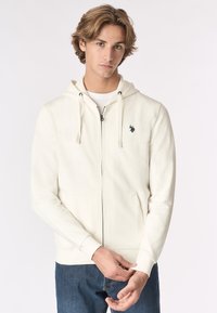 Giovane uomo con capelli castano chiaro di lunghezza media, che indossa una felpa crema con cerniera e logo, mentre sistema il polsino della manica, in piedi su uno sfondo neutro.