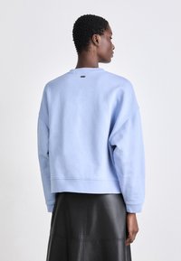 Világoskék pulóver, kerek nyakkivágással és lecsúsztatott vállakkal. Puha pamut anyag, cropped hossz, és egy kis logó részlet a hátsó oldalán.