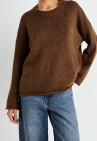 Pull en maille marron avec un col rond et une coupe ample. La texture est douce et le matériau semble chaud, complétant parfaitement un jean bleu.