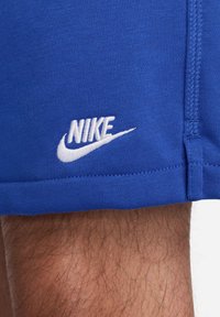 Blå Nike-shorts i lätt tyg, med en vit broderad logga på fållen och en slät, platt midjerem.