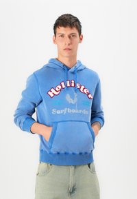 Blue cotton sweatshirt with a hood, front pocket, and ribbed cuffs. Features red and silver text "Hollister Surfboards" with graphics.