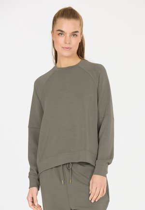 Vrouw die een bijpassende olijfgroene trui met lange mouwen en broek met trekkoord draagt, staand tegen een effen lichte achtergrond.
