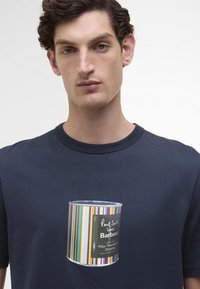 T-shirt de algodão azul-marinho com um design impresso de lata de conserva, apresentando riscas coloridas e um rótulo preto com a inscrição "Cera Impermeabilizante Thornproof".