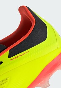 Chaussure de football jaune vif avec des accents noirs, des bords rouges et une semelle texturée. Présente la marque "HYBRIDTOUCH 2.0" sur le côté.