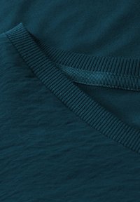 Turkusowa bawełniana tkanina o teksturowanym wykończeniu, z żeberkowym dekoltem w kształcie litery V oraz detalami wzdłuż szwu na ramieniu. Gładki, jednolity wygląd.