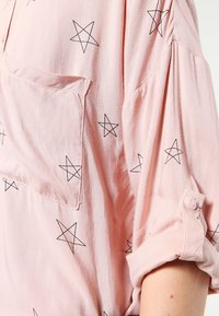 Blouse rose clair avec un motif d'étoiles noires, dotée d'une poche poitrine et de manches retroussées. Matière douce avec une coupe décontractée.