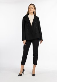 Black double-breasted coat with wide lapels, paired with black fitted trousers and high heels, set against a neutral background.