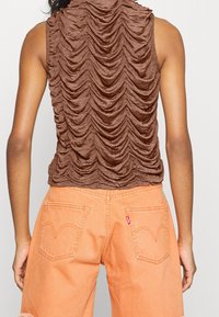 Brun texturerad ärmlös topp med horisontell rynkning, kombinerad med orange denimshorts som har bakfickor och synliga sömnadsdetaljer.