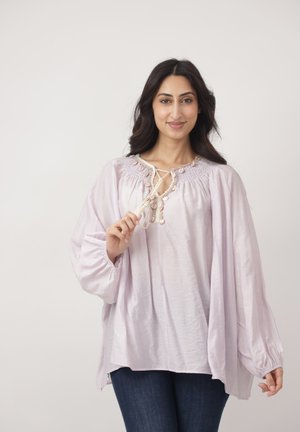 Kobieta o długich ciemnych włosach, ubrana w luźną jasnofioletową bluzkę i ciemne dżinsy, trzymająca wiązanie bluzki przy kołnierzu, uśmiechająca się do kamery.
