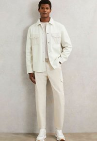 Chaqueta blanca de botones con dos bolsillos en el pecho, sobre una camisa azul claro, combinada con pantalones cargo crema y zapatillas blancas.