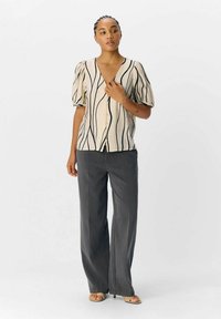 Cardigan beige à manches courtes avec des rayures noires ondulées, devant à boutons, associé à un pantalon large gris et des sandales beige clair.