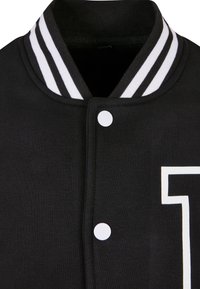 Col droit de veste universitaire noire avec doubles bandes blanches, boutons-pression à l'avant, et numéro partiel en contour blanc sur la poitrine.